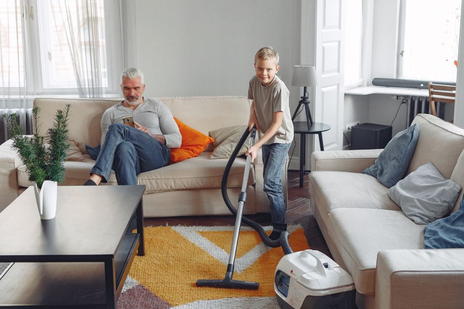 A professional cleaner wearing a full white protective suit, face mask, goggles, and blue gloves, is conducting a deep surface cleaning on a beige carpet with a steam cleaning machine, generating stea